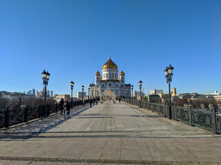 Fototapeta premium Moscow, Russia - March 01, 2022: View of the Cathedral of Christ the Savior from the Patriarchal pedestrian bridge against the blue sky. People are walking on the bridge