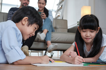 Asian Thai siblings are lying on living room floor, drawing homework with colored pencils together, parents leisurely relax on a sofa, lovely happy weekend activity, and domestic wellbeing lifestyle.