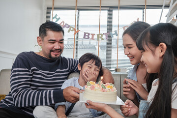 Happy Asian Thai family, young son is surprised with birthday cake, blows out candle, and celebrates joy party with parents and sister in living room together, wellbeing domestic home special event.