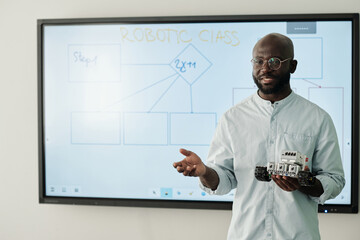 Young teacher of robotic class holding new model of toy robot at lesson while standing by...