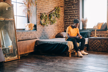 Attractive man texting on his mobile phone sitting on the bed indoors at home