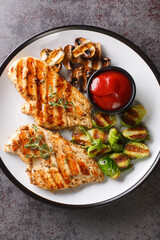 Grilled chicken fillet with Brussels sprouts, mushrooms and spicy tomato sauce close-up in a plate on the table. Vertical top view from above