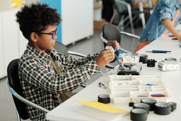 Clever African American schoolboy constructing new robot by desk at lesson of robotics and...
