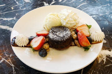 A plate of chocolate cake with vanilla ice cream, berry, and strawberry on a black marble table.