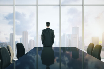 Back view of young businessman standing in modern meeting room interior with reflections on table, chairs and panoramic window with sky clouds view and daylight. CEO and conference concept.