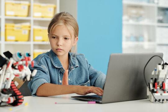 Cute blond schoolgirl looking at toy robot while sitting by desk in front of laptop and entering description of new creation at lesson