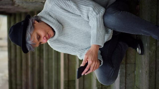 Portrait of mature woman sitting on stairs, using smart phone and looking into camera.
