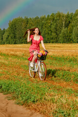 A young beautiful woman rides a bicycle across the field against the backdrop of a forest and a rainbow in the sky and enjoys with the sun