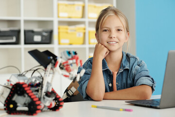Cute blond schoolgirl looking at camera with smile while sitting by desk in front of laptop and handmade robot constructed at lesson