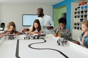 Young teacher of robotics with tablet helping schoolkids control their robots during play after...