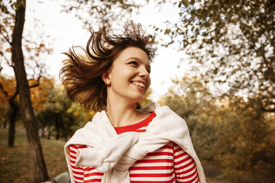 Close up of beaming young caucasian girl laughing looking to side outdoors. Her dark short hair flutters in breeze, wearing sweater. Lifestyle, female beauty concept