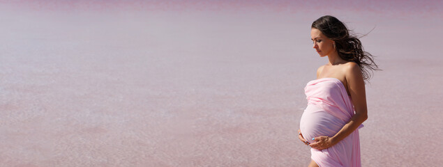 Pregnant woman stands on sea beach in summer in white light dress with fluffy hair in the wind....