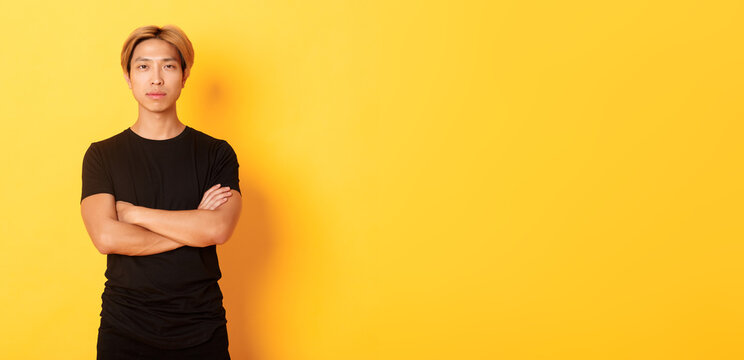 Portrait of serious-looking confident asian guy in black t-shirt, cross arms chest, standing yellow background