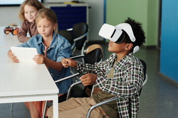 Side view of cute African American schoolboy in vr headset controling virtual robot while his classmate watching action on tablet screen