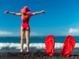 Red flip flops in the black sand and silhouette of woman in red swimsuit standing on the ocean beach. Summer concept.