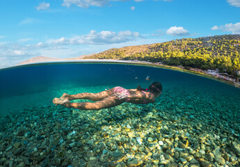 Girl diver is snorkeling on a beautiful sea beach. The lower half of the image is occupied by the seabed, the upper half by the coast and the sky.