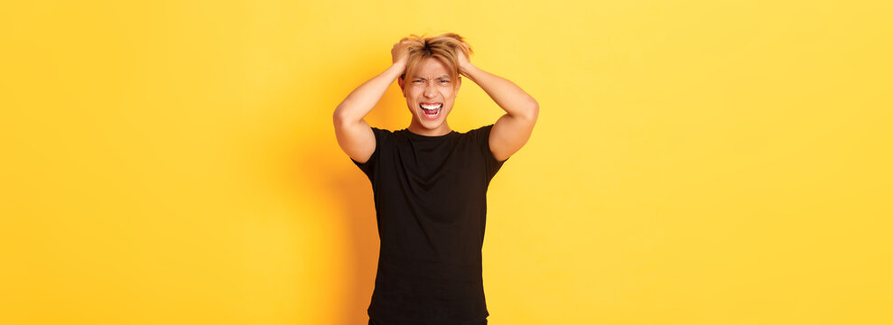 Portrait of pissed-off angry asian man tossing haircut and yelling furious, standing over yellow background
