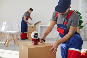 Moving company worker in uniform packing stuff, holding gun roller dispenser and sealing full...