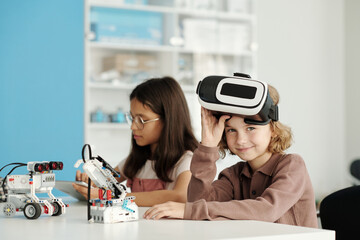 Happy youthful schoolchild taking off vr headset and looking at camera while sitting by desk next...