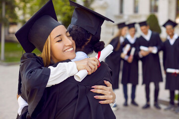 Fototapeta Positive women graduates in student gown hug each other after receiving university or college diploma and rejoice at completion of their education standing under open sky. Selective background obraz