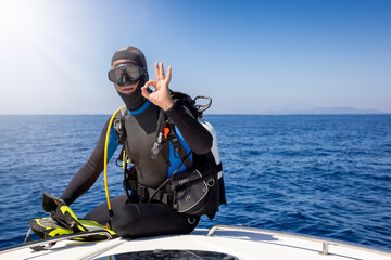A scuba diver in full gear sits on a boat and signals the OK sign