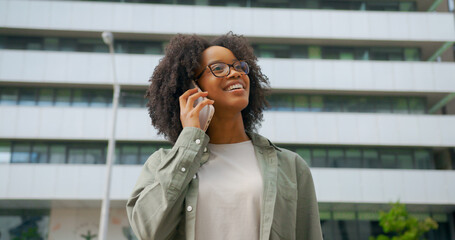 Fototapeta premium Portrait of cheerful mixed-race curly haired woman having a call by her phone outdoors, wearing casual cloth