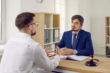 Young man talking to a professional lawyer. Serious attorney in a formal suit sitting at his office desk and listening to a client telling about his problem. Law consultation, legal advice concept