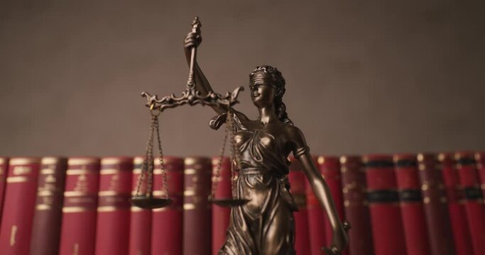 bronze blindfolded goddess representing law statue spinning, holding a scale and sword symbolizing impartiality and two broad sources of law in a university room with a background made of books