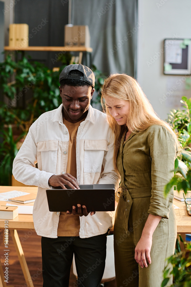 Wall mural two young intercultural employees in casualwear looking at laptop screen while standing by desk and 