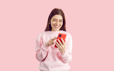 Online connectivity, working or socializing remotely. Joyful young woman on pastel pink background swipes and scrolls on screen of mobile phone. Smiling woman smiling looking at smartphone screen.