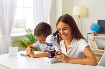 Children and science. Schoolgirl is looking into microscope at home with her younger brother, preparing for science lesson. Funny children sitting at table with microscope and writing in notebook.