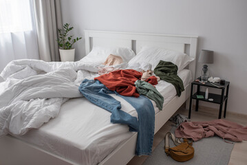 Clothes thrown all around the bed. Cluttered bedroom with a white bedding and a minimalistic modern interior.