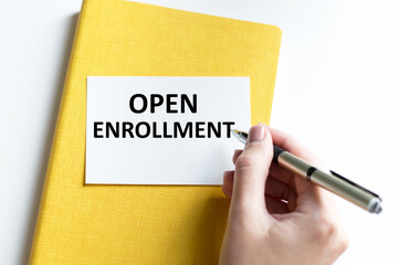 Card with text Open Enrollment on the background of a notebook on the table next to the hand of the man holding a pen
