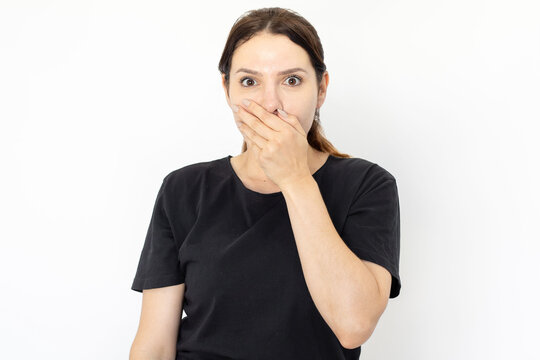 Portrait of surprised young woman. Female model in black t-shirt with dark hair looking at camera, covering mouth with hand. Portrait, studio shot, surprise concept