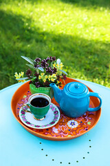 A green coffee cup, a bouquet of wild flowers and a turquoise coffee pot stand on an orange tray. The tray stands on a turquoise table in a sunny garden
