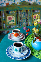 A still life with two coffee cups on a turquoise kitchen table. On the table is a vase with flowers. In the background you see a beautiful Indian wooden sofa. It's a colorful photo.