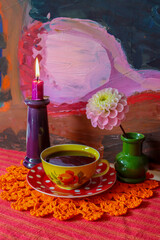 A still life with a yellow coffee cup standing on an orange lace cloth. Next to it is a candlestick...