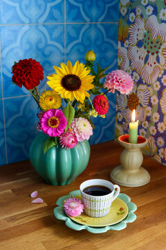 A cup of coffee in the kitchen. In the picture there is also a lighted candle and a green vase with sunflowers and dahlias. In the background, turquoise Moroccan tiles and a blue floral wallpaper. 