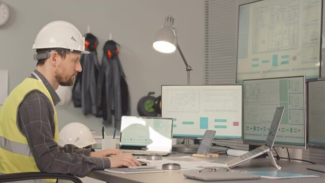 Waist up slowmo of male engineer in safety uniform sitting by computer at office desk developing details for industrial equipment