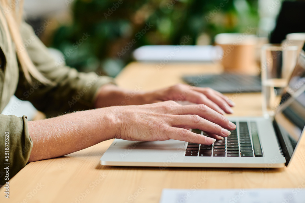 Poster side view of hands of young businesswoman typing on laptop keyboard while sitting by table in office
