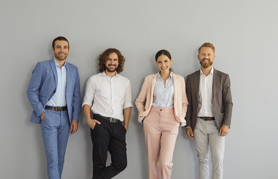 Successful Stylish Business Team Standing In Studio. Group Portrait Of Four Happy Confident Young People In Smart Office Suits Standing Together Against Grey Wall, Looking At Camera And Smiling