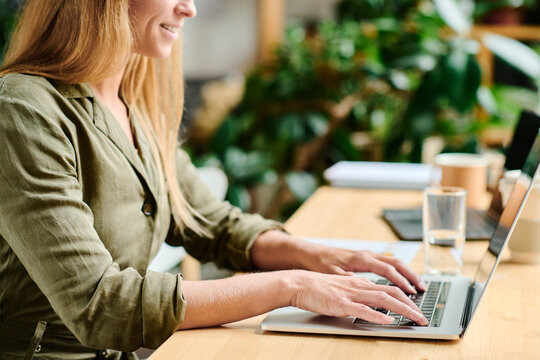 Close-up of young female freelancer in casualwear touching buttons of laptop keypad while sitting by table in cafe or modern office