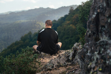 Naklejka premium a strong athletic man sits on a high mountain rise with his back to us, rests and enjoys the view of nature during a hiking trip
