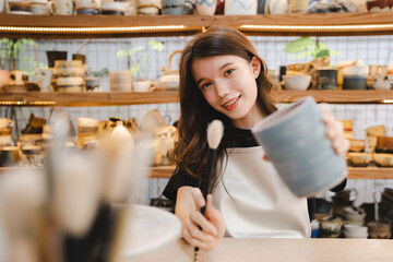 Beautiful young woman holding pottery instrument for scraping, smoothing, shaping and sculpting. Lady siting on bench with pottery wheel and making clay pot