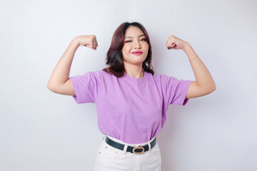 Excited Asian woman wearing a lilac purple t-shirt showing strong gesture by lifting her arms and muscles smiling proudly