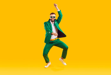Fototapeta na wymiar Cheerful energetic man has fun dancing in gangnam style at St. Patrick's Day party. Full length of man in trendy green suit and sunglasses of same color doing funny dance moves on orange background.