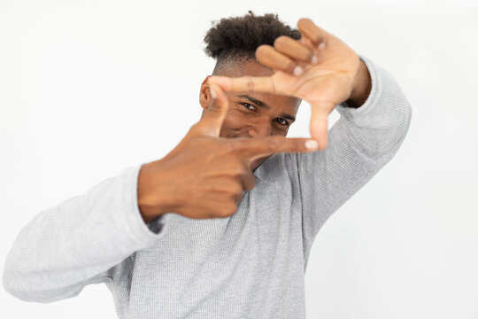 Portrait of happy African American man making frame gesture. Young bearded guy wearing white sweater looking at camera in finger frame against white background. Photographing concept