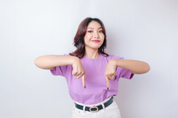 Fototapeta na wymiar Excited Asian woman wearing lilac purple t-shirt pointing at the copy space downside her, isolated by white background