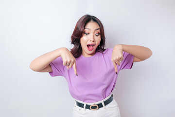 Shocked Asian woman wearing lilac purple t-shirt pointing at the copy space downside her, isolated by white background