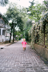 Fototapeta na wymiar Little girl runs along the paving stones past the wrought-iron fence of the park. High quality photo